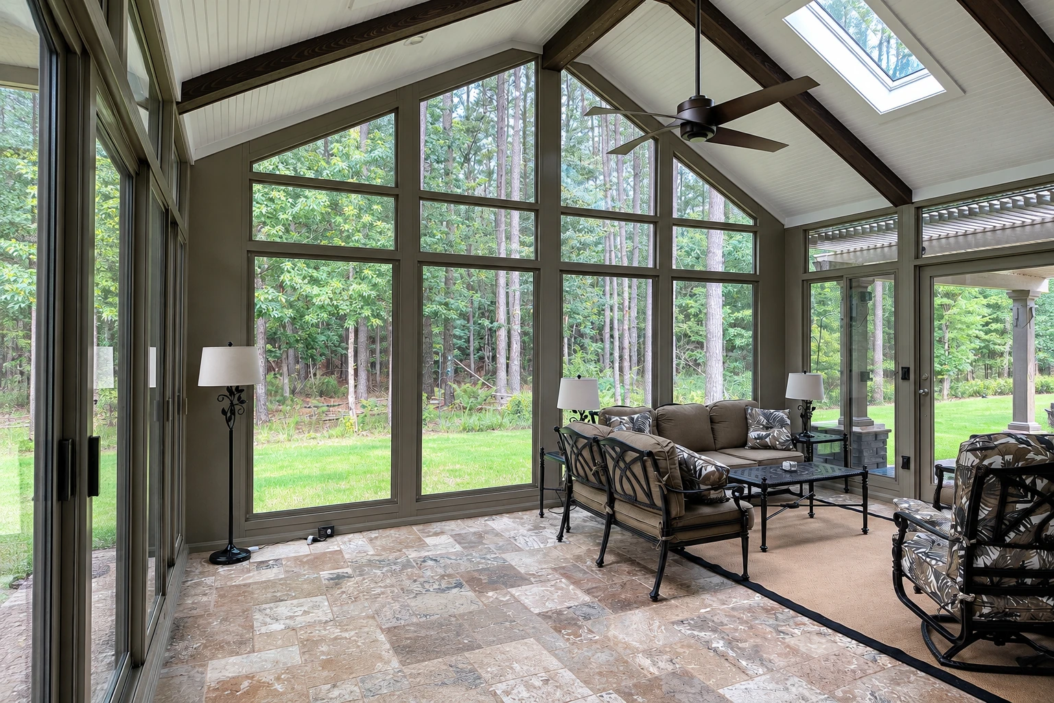 An all-season sunroom has tall windows overlooking a wooded lawn, a vaulted white ceiling with dark beams, and a furnished seating area.