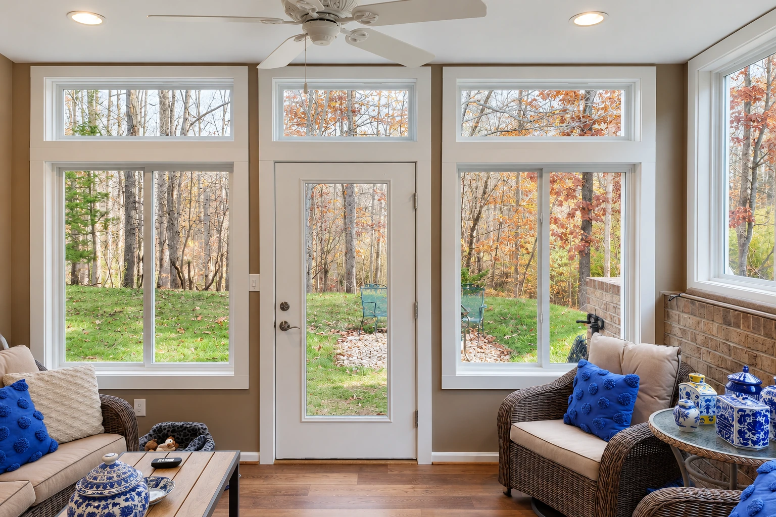 A sunroom interior features a glass-panel door, large windows, wicker seating and views of autumn trees.