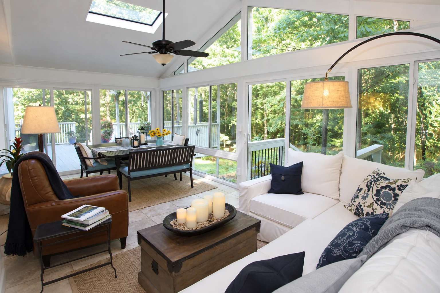 A bright glass sunroom with a high ceiling and skylight combines a dining area with white sofas and a wood coffee table.