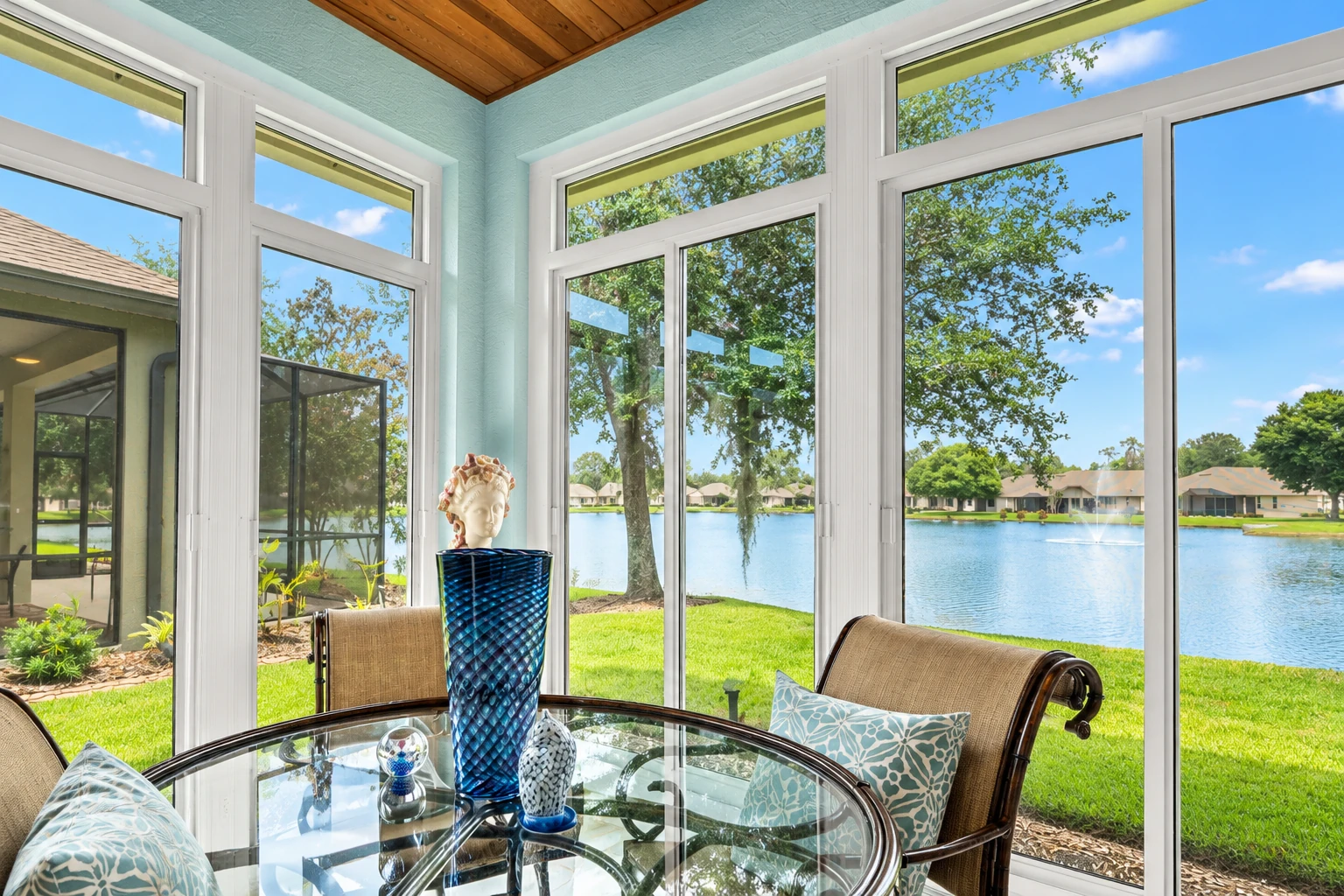 A bright sunroom with white-framed windows overlooks a lake, with a glass table and cushioned chairs in the foreground.