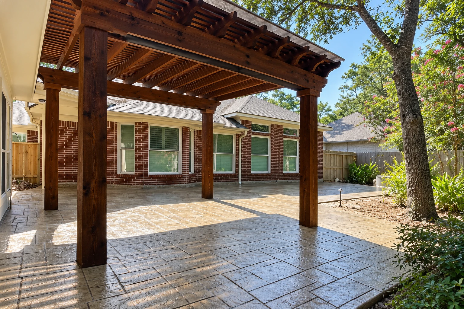 A dark-stained wooden patio cover shelters a broad paved backyard patio beside a brick house and mature trees.