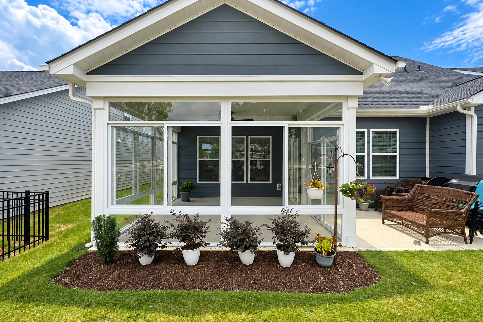 A glass-enclosed patio sunroom with a peaked roof adjoins a dark blue house and outdoor seating area.