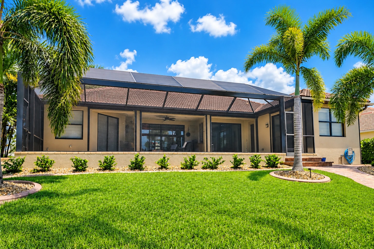 A screened patio enclosure with a dark frame and roof spans the back of a house, bordered by palm trees and a green lawn.