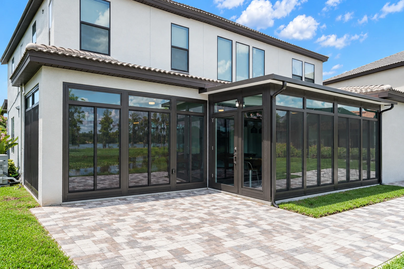 Sunroom addition with dark-framed floor-to-ceiling windows attached to a white two-story home, opening onto a paved patio.