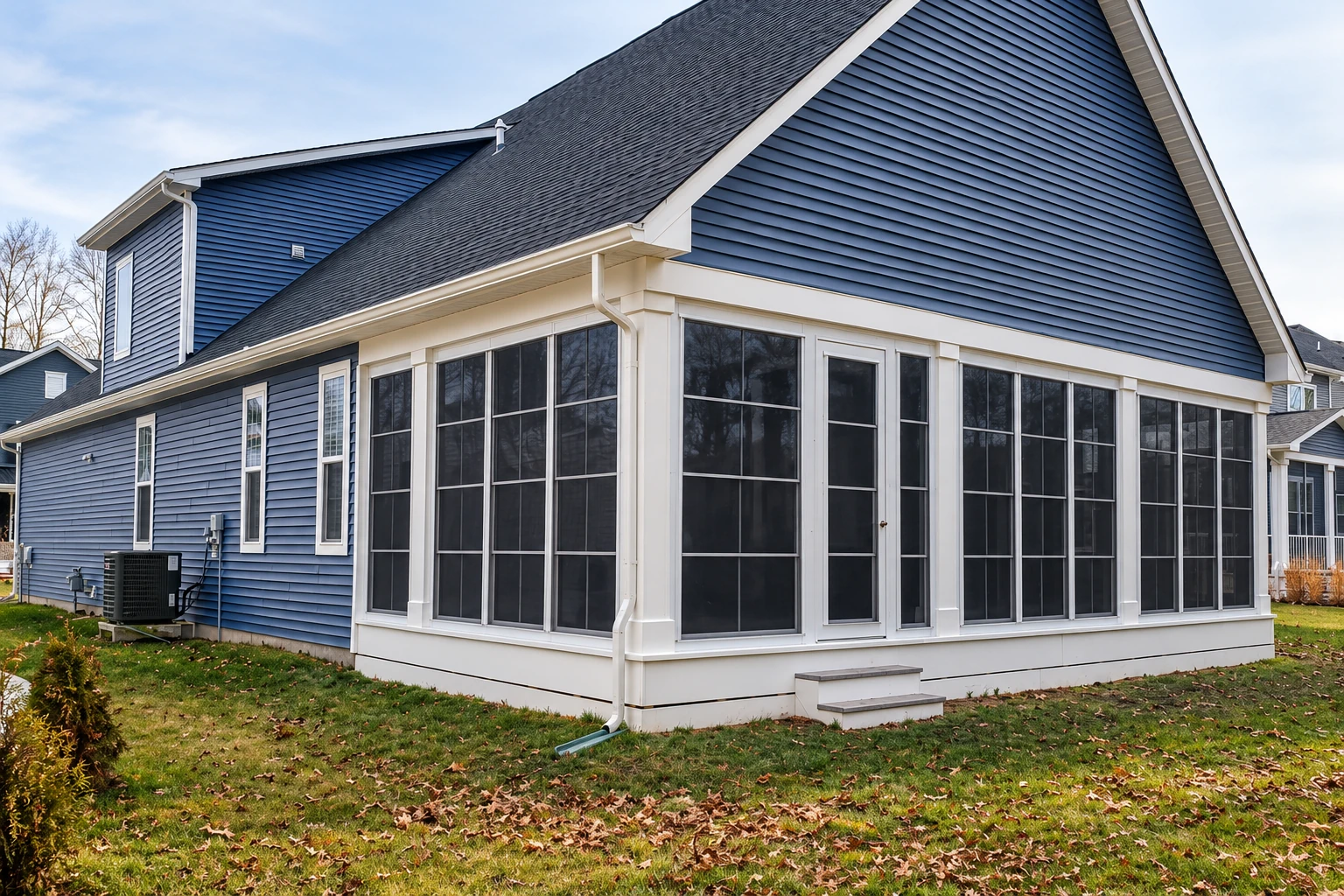 A large sunroom addition with white framing and rows of screened windows extends from a blue-sided house above a leaf-strewn lawn.