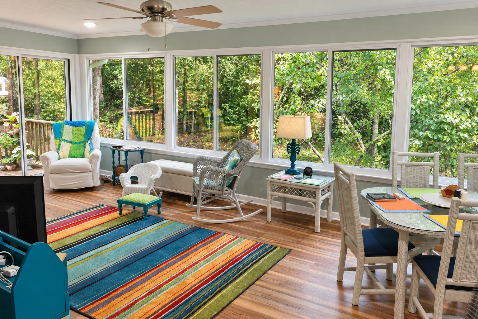 A sunroom with long windows overlooking dense greenery has wicker seating, a dining table, and a colorful striped rug.