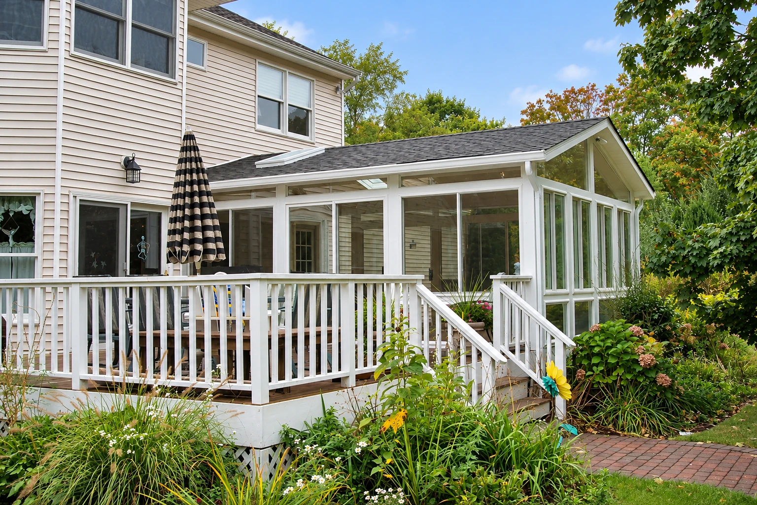 A white-framed three-season sunroom extends from a house behind a raised deck with railings and garden beds.