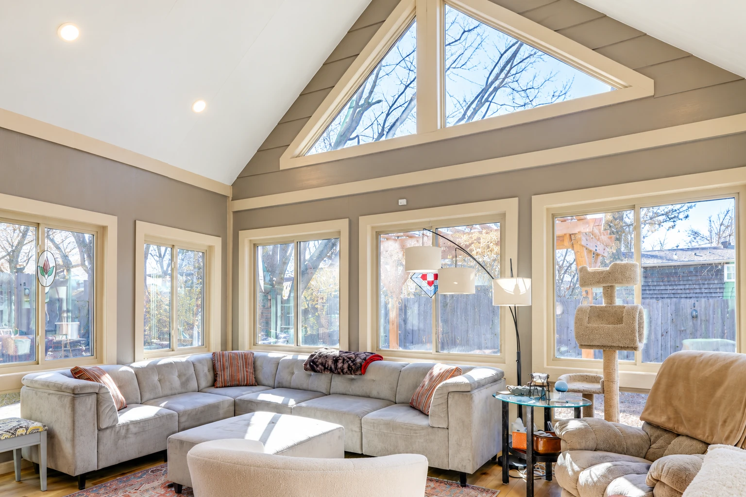 A bright sunroom with white vinyl-framed windows has a vaulted ceiling, triangular clerestory windows, and a large sectional sofa.